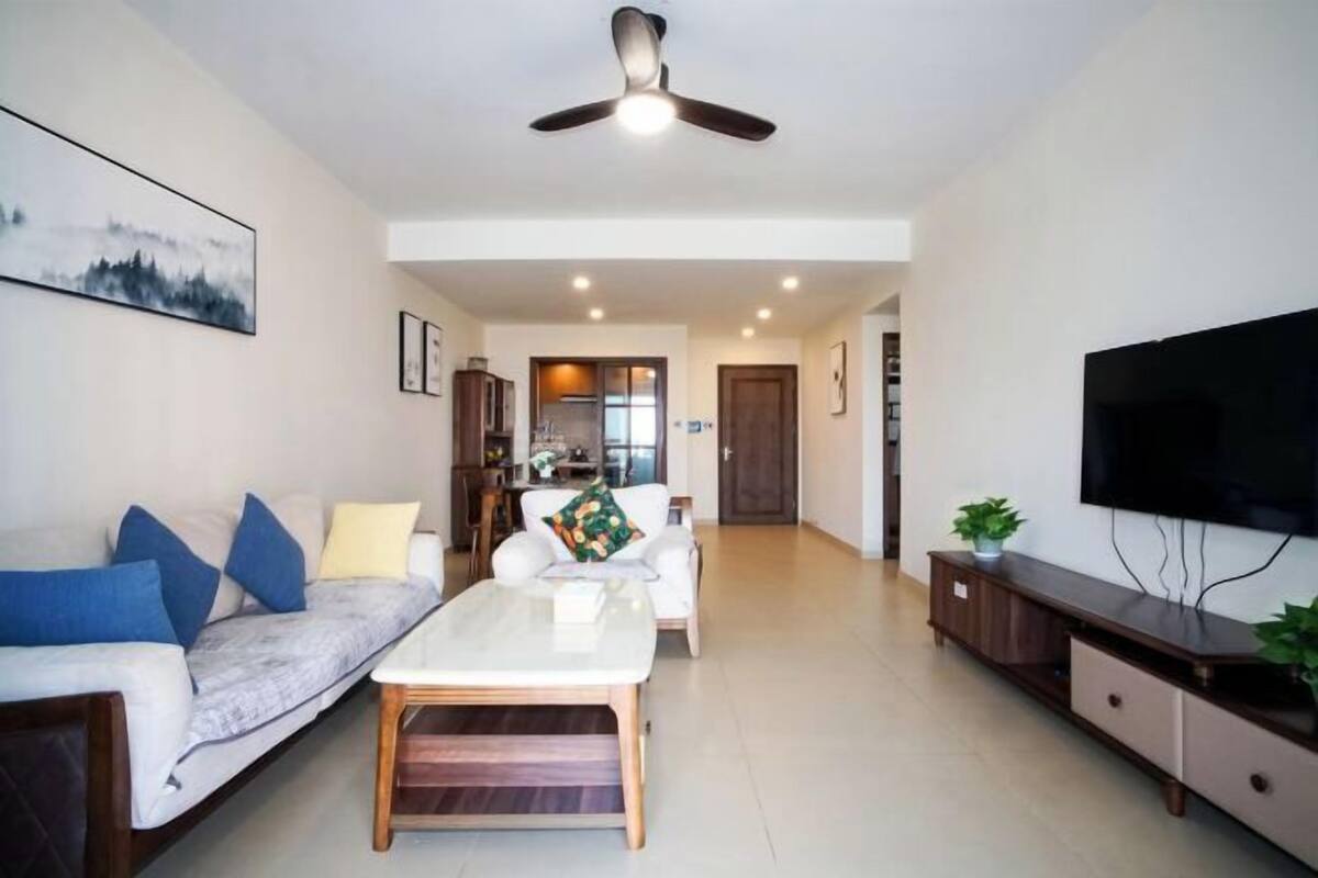 A living room with a sofa, coffee table, TV, ceiling fan, and a kitchen in the background.