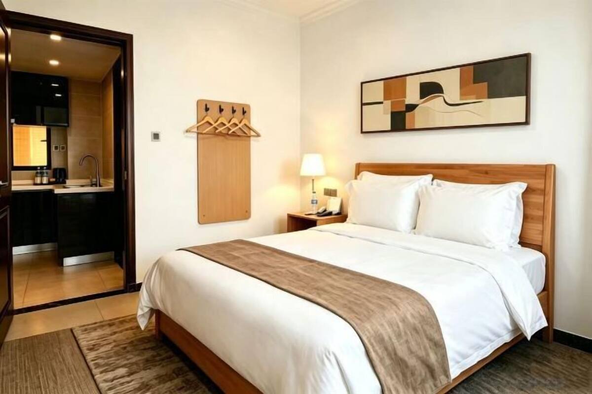 A hotel room with a bed, a wooden headboard, a nightstand with a lamp, a wall-mounted coat rack, and a painting.