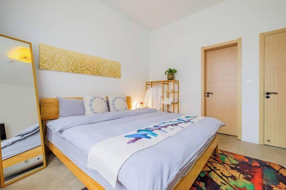 A bedroom with a bed, a bedside table, a mirror, a wall art piece, a door, and a small plant.