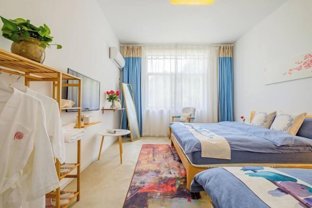 A bedroom with two beds, a wooden bedside table, a small round table, a chair, a potted plant, and a painting on the wall.