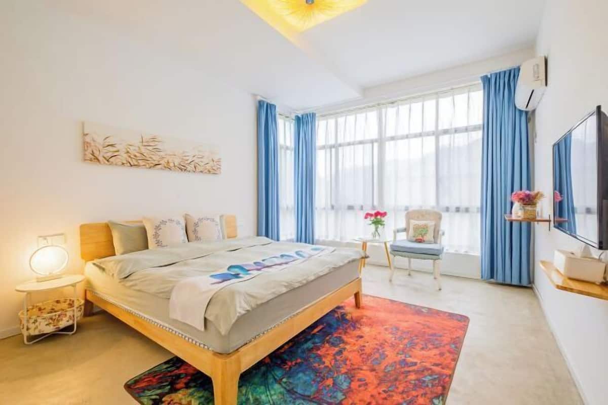 A modern bedroom with a large bed, a bedside table with a lamp, a chair, a small table with flowers, and a TV on the wall.