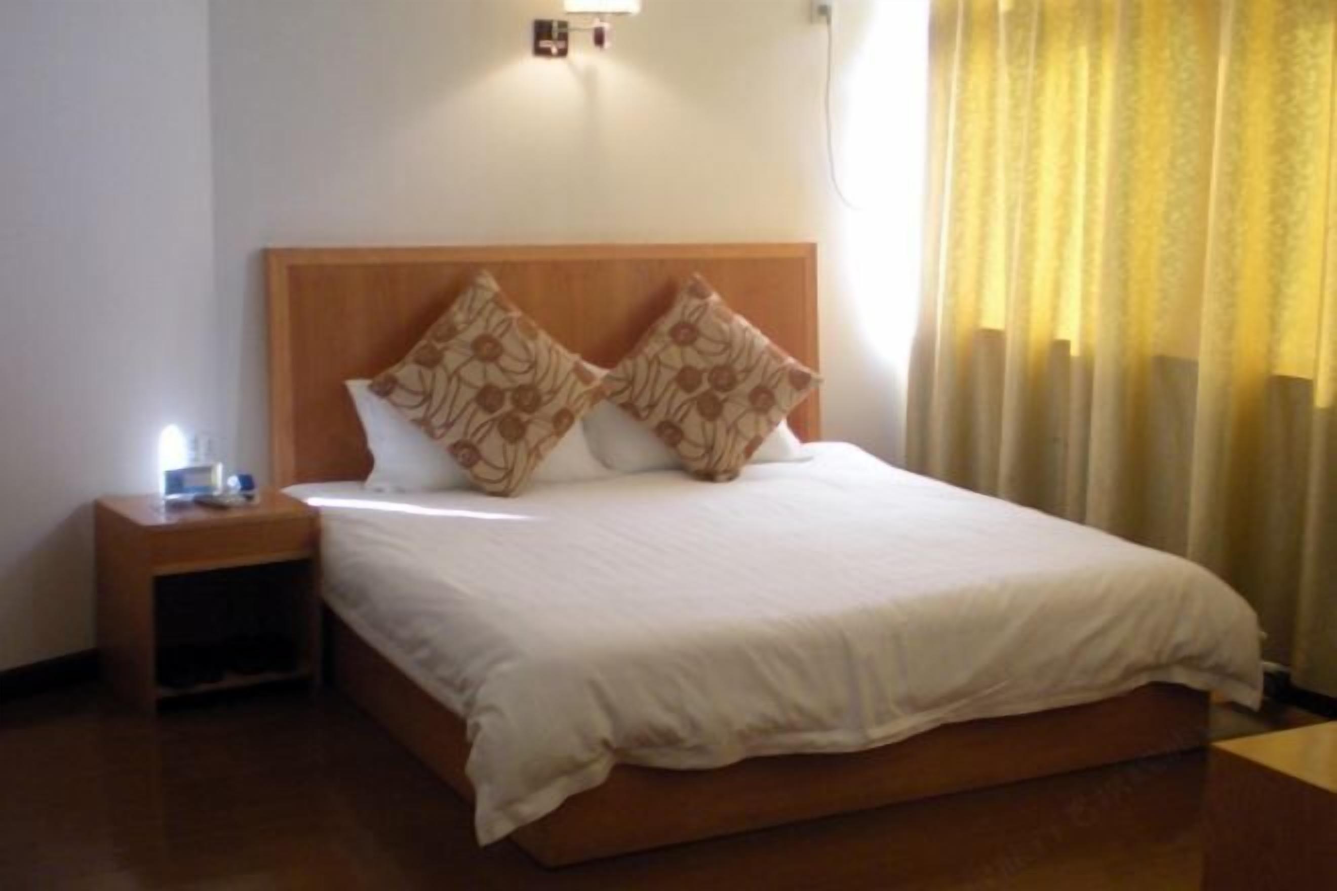 A hotel room with a wooden headboard, two patterned pillows, a white bedspread, a nightstand with a lamp, and a window with yellow curtains.