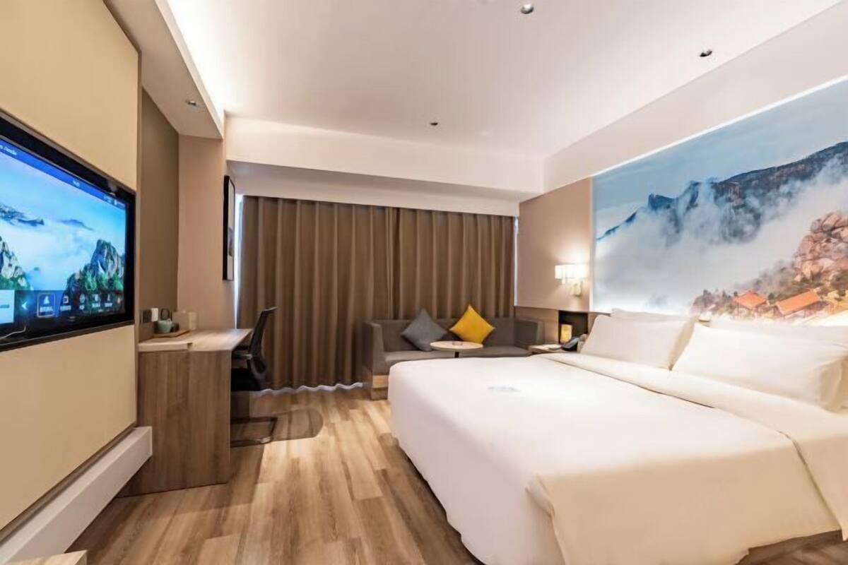 A hotel room with a large bed, a desk, a TV, and a painting of a mountain landscape.