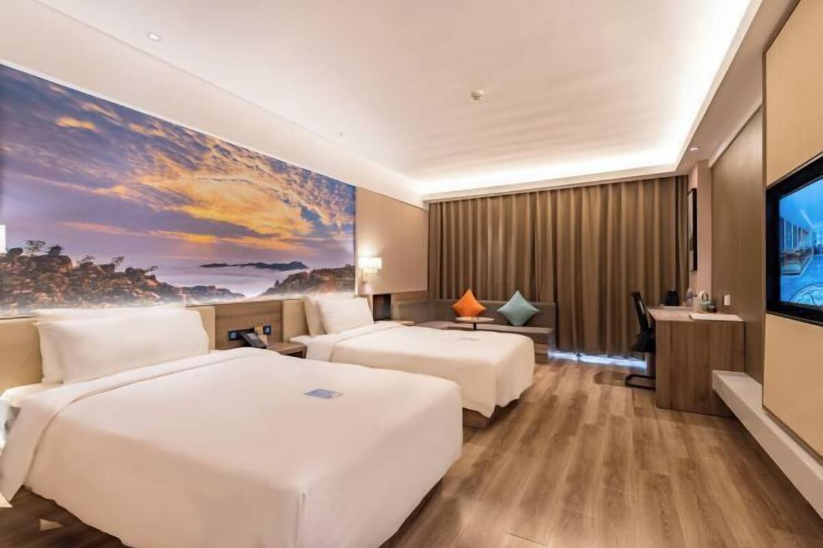 A hotel room with two beds, a desk, a TV, and a large mural of a sunset.