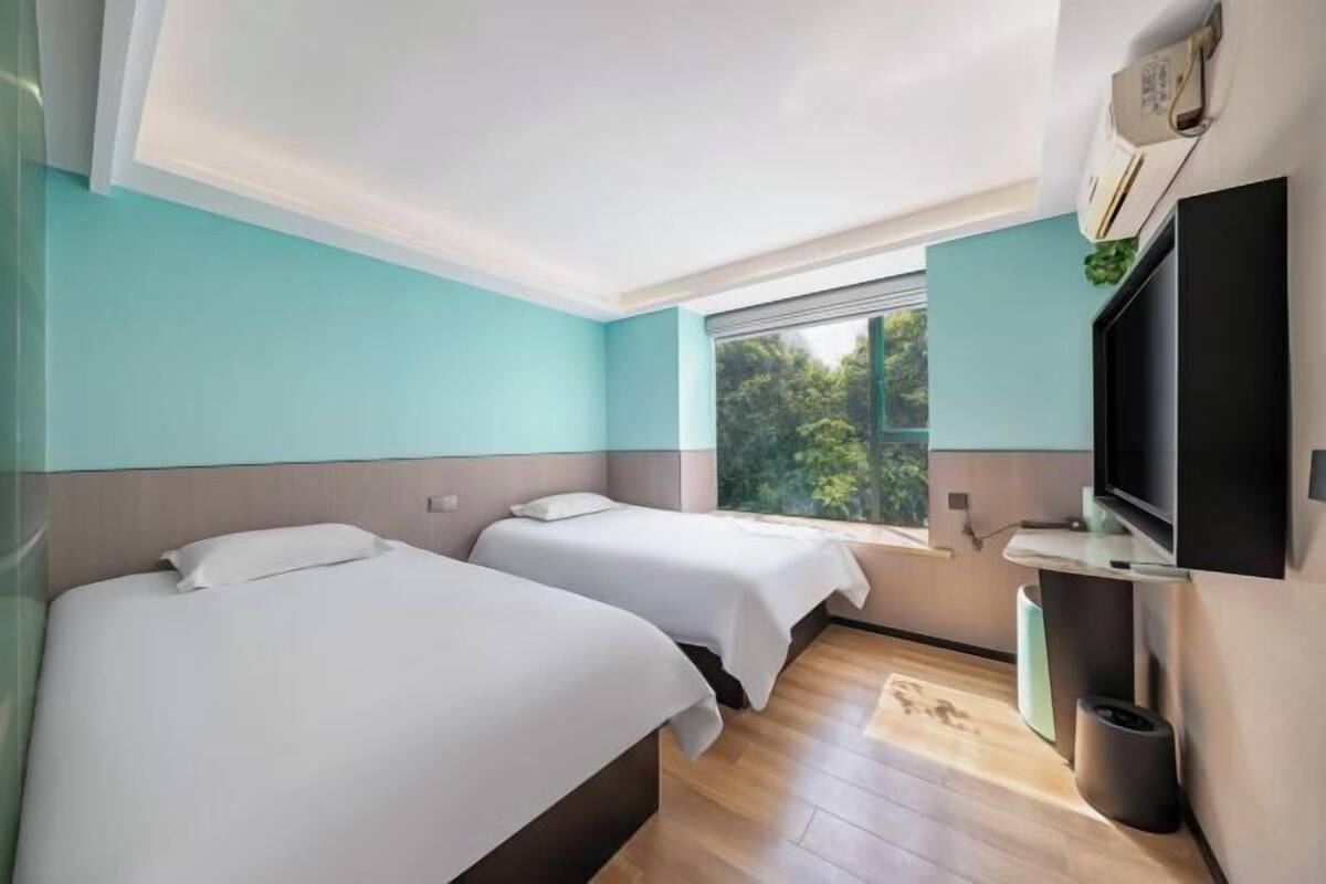 A hotel room with two beds, a TV, a window with a view of greenery, and a ceiling light.
