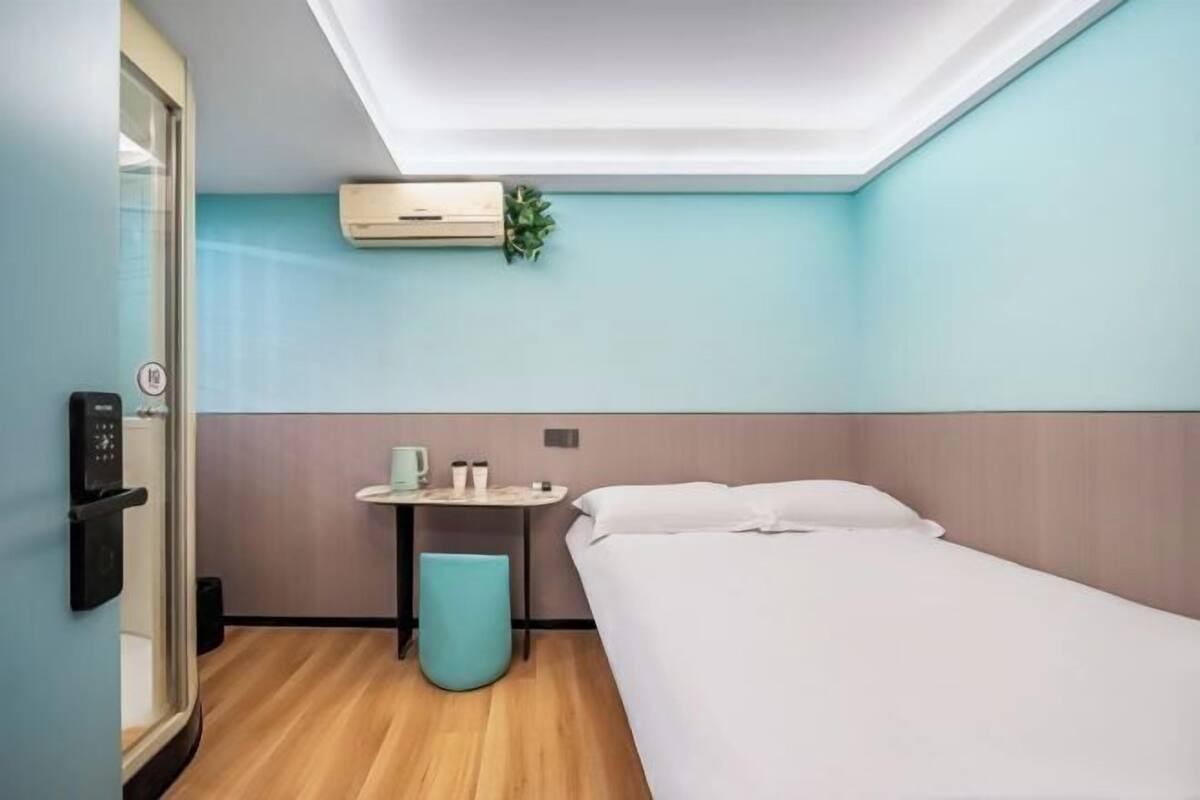 A modern hotel room with a bed, a small table with a cup and a plant, and a wall-mounted air conditioner.