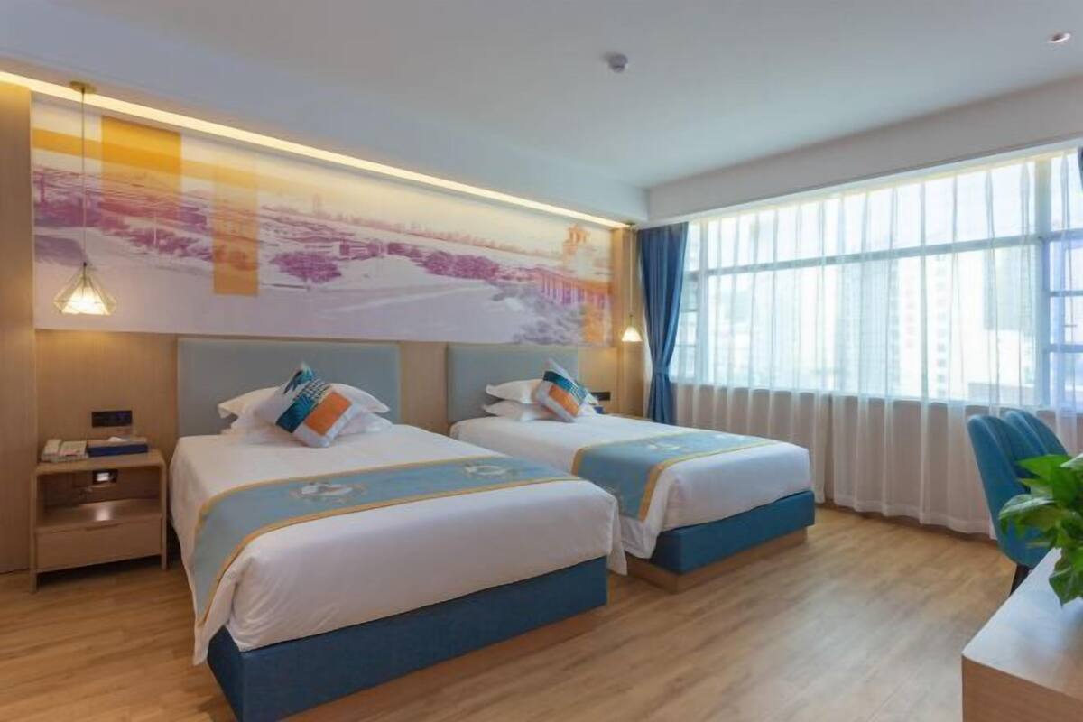 Een hotelkamer met twee bedden, een groot raam met gordijnen, een bureau met stoel en een kunstwerk aan de muur.