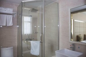 Zona de ducha con paredes de vidrio, una toalla blanca colgada en la puerta, un lavamanos blanco y un espejo sobre él.