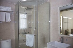 Zona de ducha con paredes de vidrio, una toalla blanca colgada en la puerta, un lavamanos blanco y un espejo sobre él.