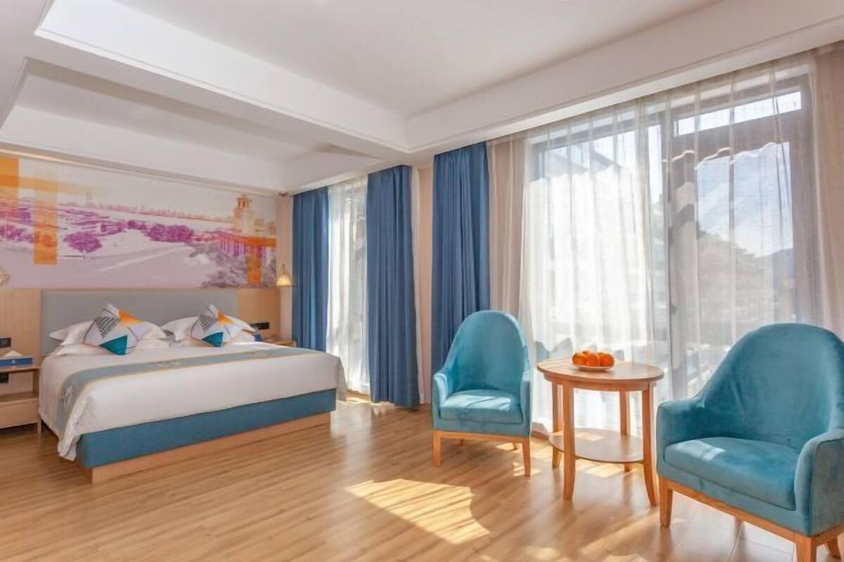 Een hotelkamer met een bed, twee fauteuils, een klein tafeltje en een groot raam met gordijnen.