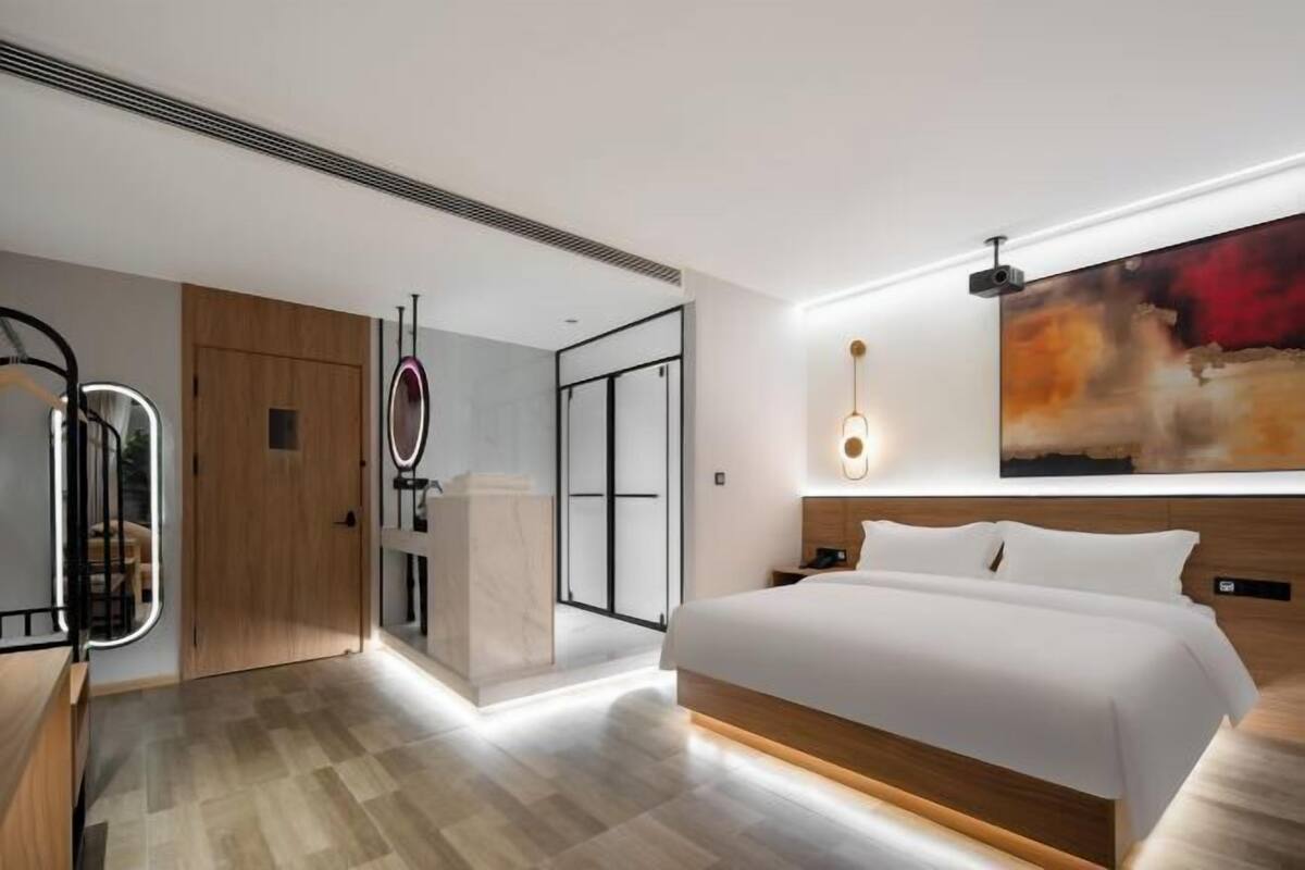 A modern hotel room with a large bed, a wooden wardrobe, a vanity with a mirror, and a large abstract painting on the wall.