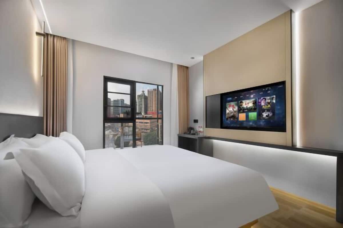 A modern hotel room with a large bed, a TV mounted on the wall, and a city view through the window.