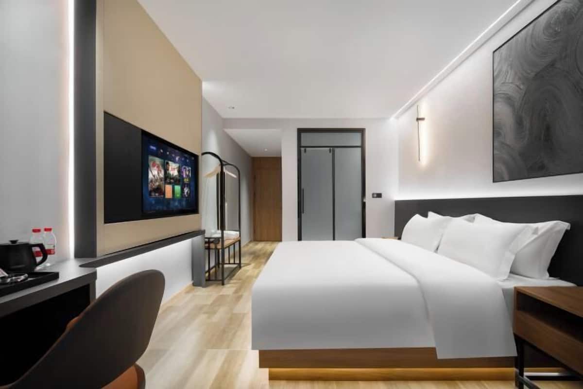 A modern hotel room with a large bed, a desk with a computer, a TV mounted on the wall, and a wardrobe.
