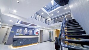 A modern hotel lobby with a reception desk, a staircase, and a high ceiling with skylights.