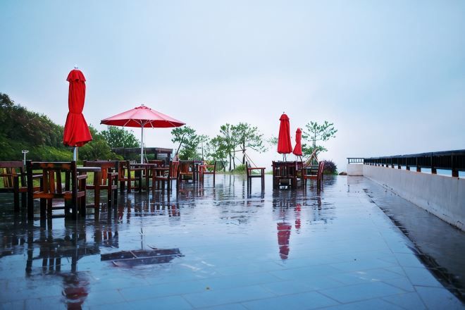 An outdoor seating area with tables and chairs, some with red umbrellas, on a wet surface.