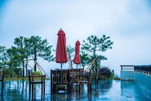 An outdoor seating area with tables and red umbrellas, surrounded by trees and water. - Green Field Manor (Ningbo)