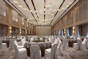 A banquet hall with round tables, white chairs, chandeliers, and a stone wall.