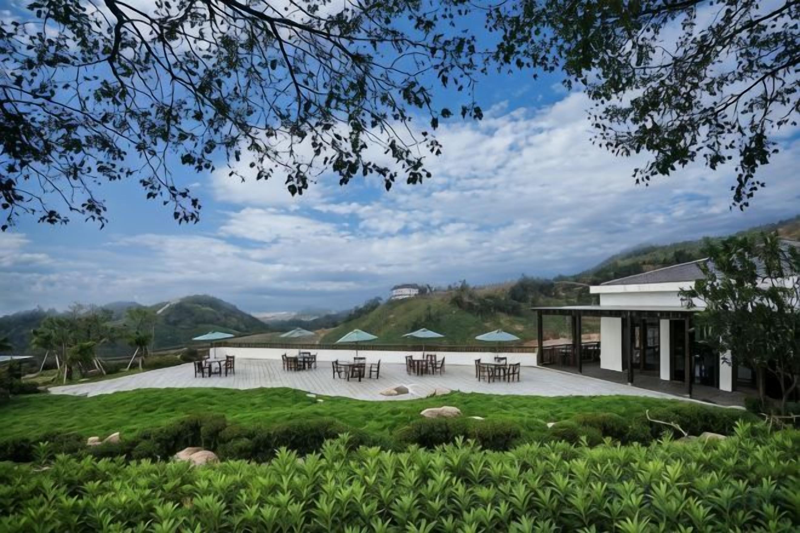 An outdoor seating area with tables and chairs, surrounded by greenery and hills in the background.