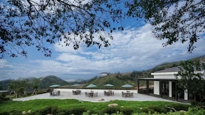 An outdoor seating area with tables and chairs, surrounded by greenery and hills in the background. - Green Field Manor (Ningbo)
