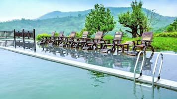 A pool with wooden chairs and a metal railing, surrounded by greenery and hills.