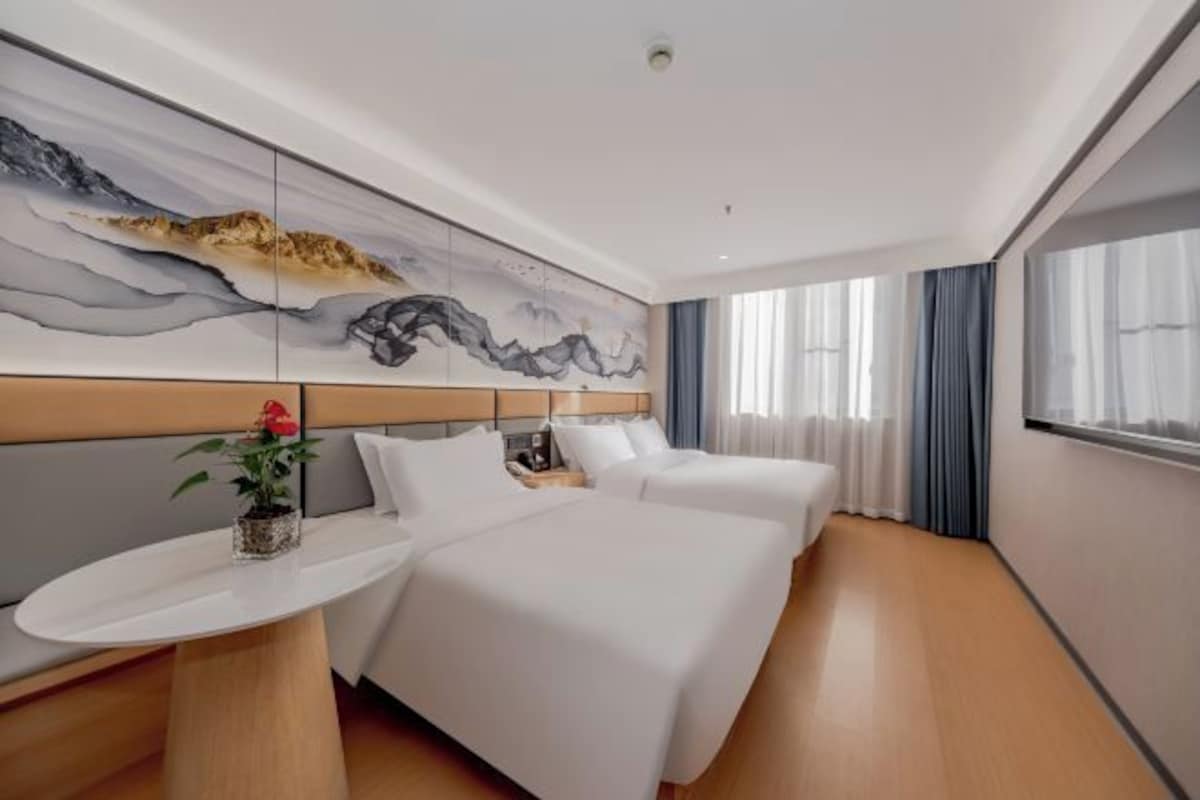 A hotel room with two beds, a large wall mural, a bedside table with a vase of flowers, and a window with curtains.
