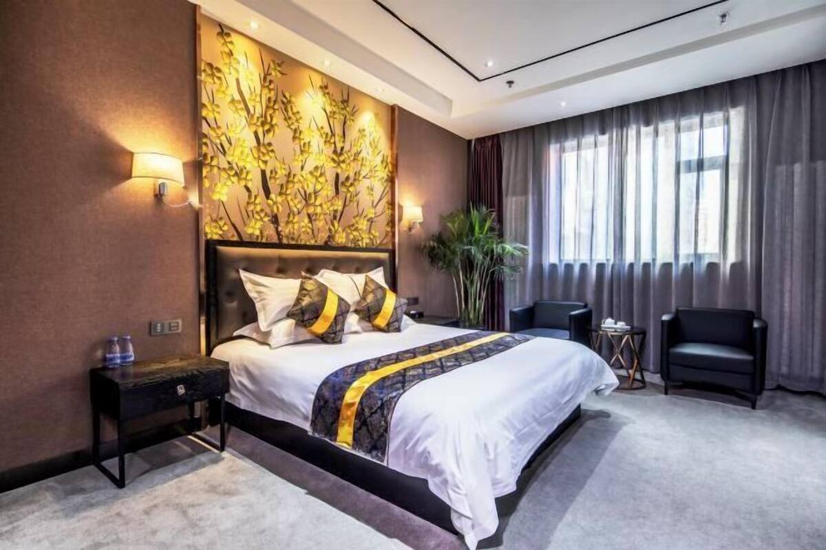 A modern hotel room with a large bed, a headboard with a decorative wall piece, a side table, a chair, and a large window with curtains.