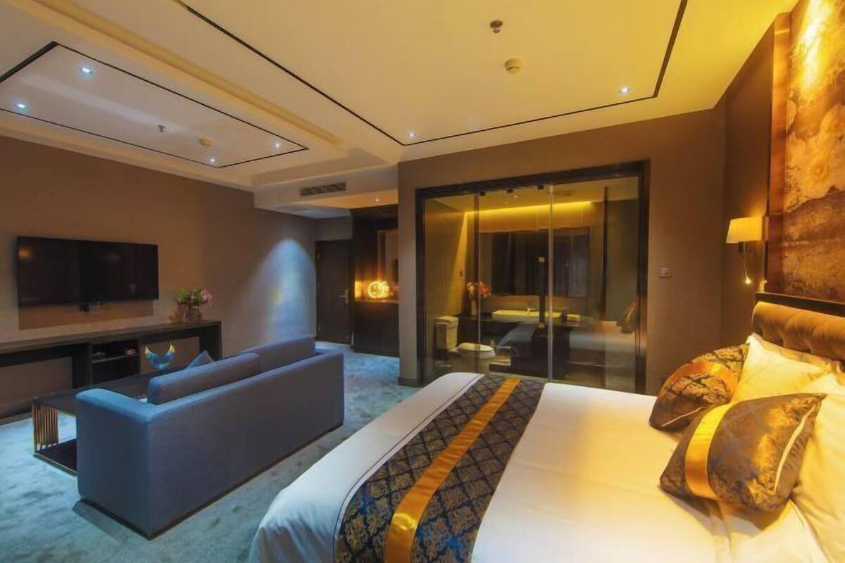 A modern hotel room with a large bed, a sofa, a TV, and a glass-enclosed bathroom.