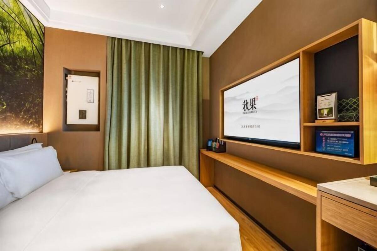 Une chambre d’hôtel avec un lit, une télévision, une étagère avec des livres et une fenêtre avec des rideaux.