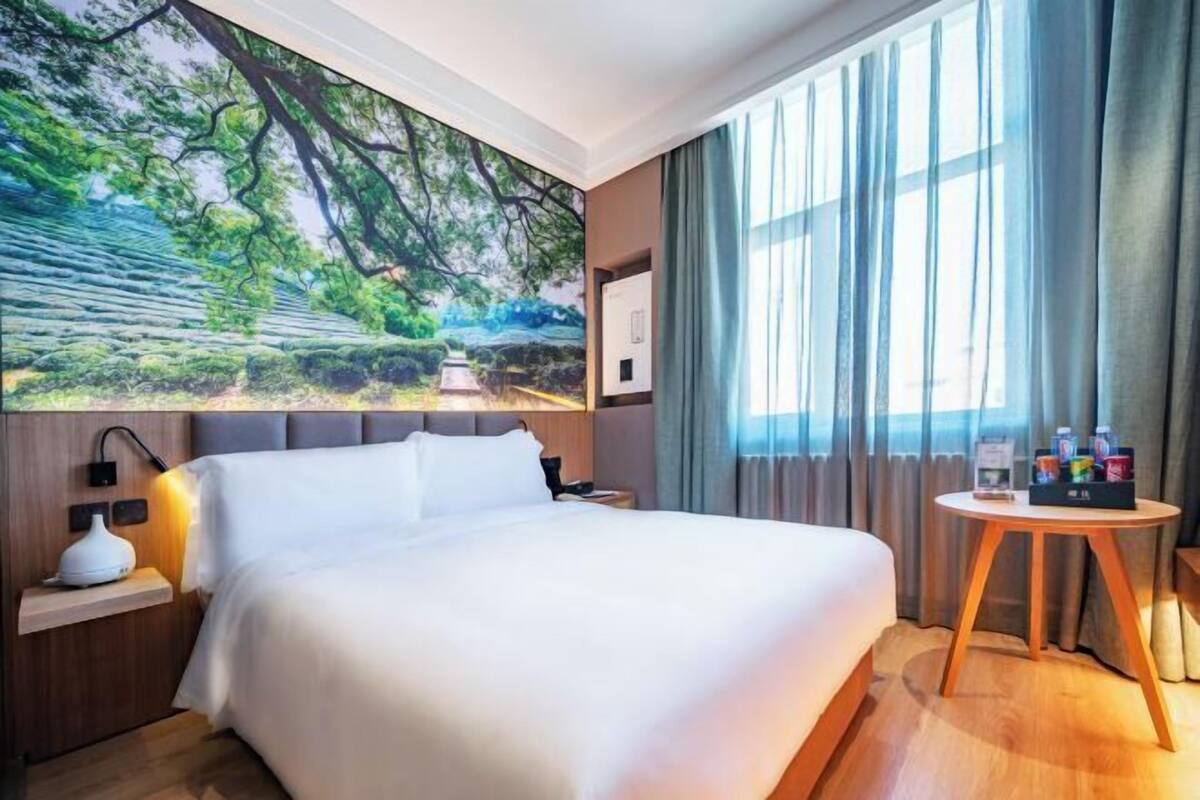 Une chambre d’hôtel avec un grand lit, une table de chevet, une lampe fixée au mur, une petite table ronde avec des articles de toilette et une grande fenêtre avec des rideaux.