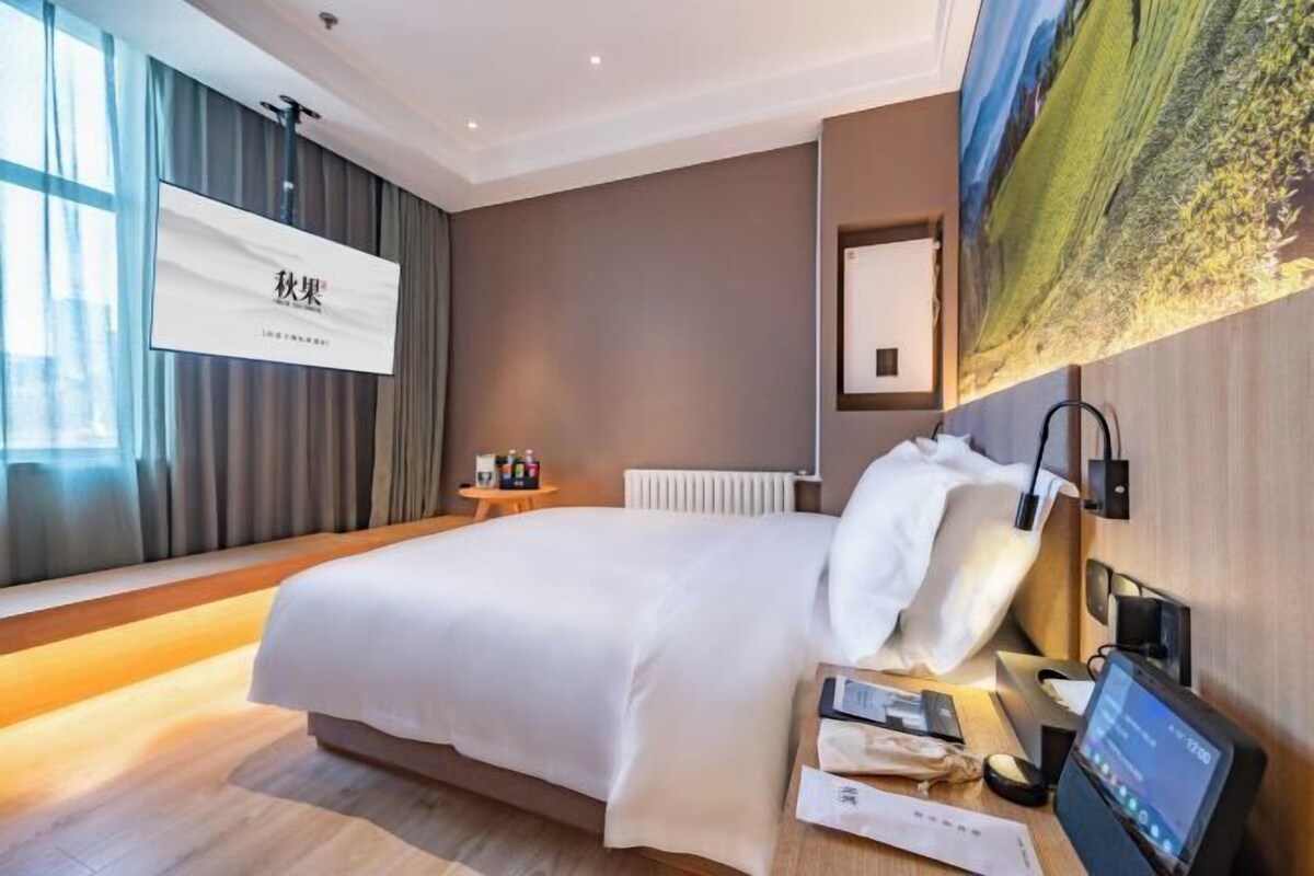 A modern hotel room with a large bed, a bedside table with a digital device, and a view of a landscape painting on the wall.