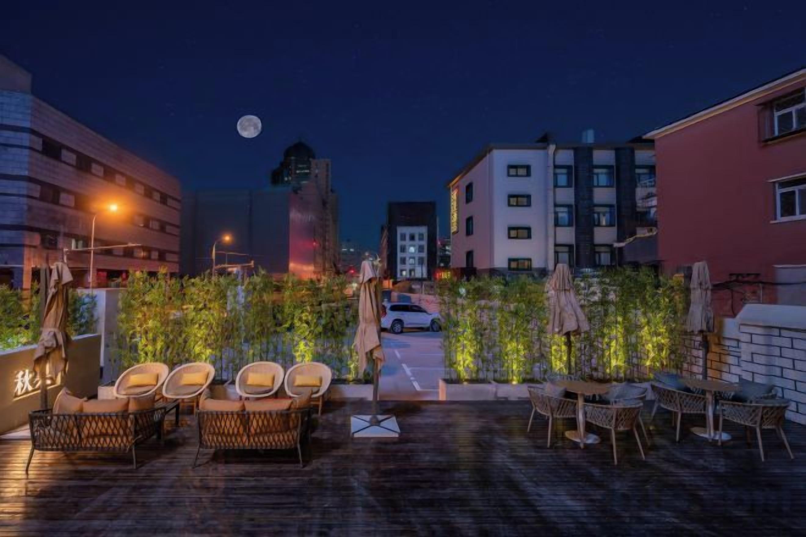 Uma área externa com mesas e cadeiras de vime, guarda-sóis e piso pavimentado, com edifícios ao fundo.