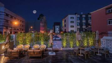 Uma área externa com mesas e cadeiras de vime, guarda-sóis e piso pavimentado, com edifícios ao fundo.