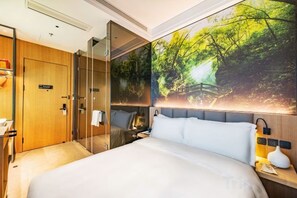 Habitación de hotel con una cama grande, una pared de vidrio, una puerta de madera y una lámpara fijada a la pared.