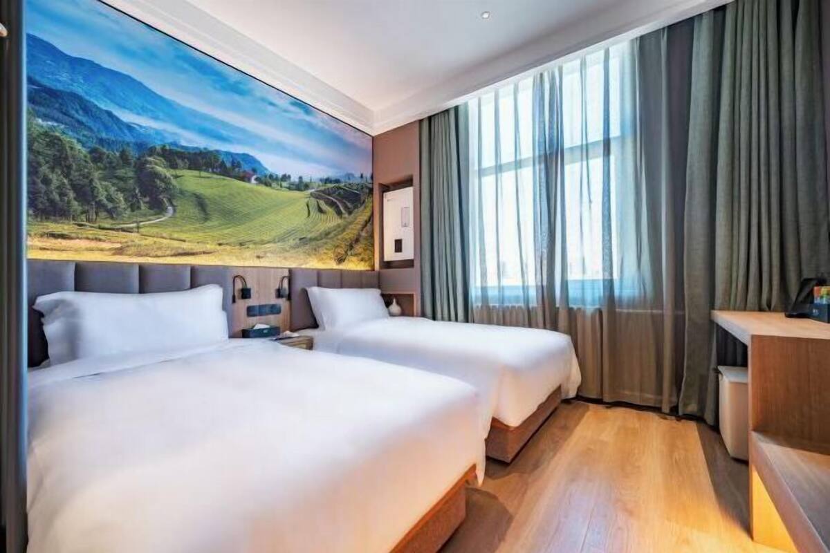 Une chambre d’hôtel avec deux lits, une grande fenêtre avec des rideaux, un tableau représentant un paysage et un bureau avec une chaise.