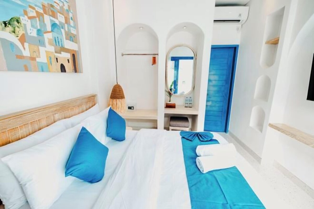 Una habitación de hotel con cama, decoración azul y blanca, cabecero de madera, espejo montado en la pared y puerta azul.