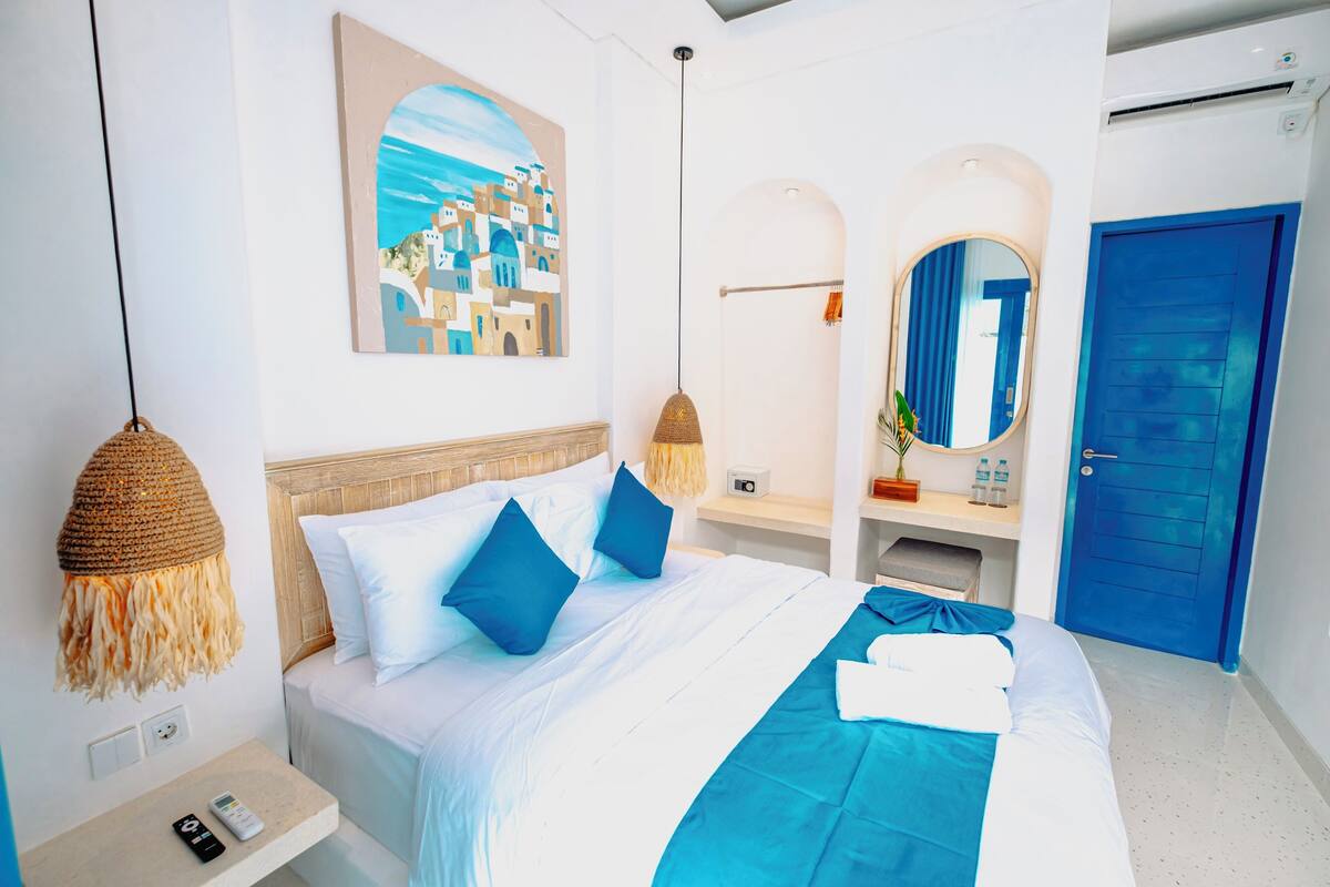A hotel room with a bed, a nightstand, a wall-mounted painting, a mirror, and a blue door.