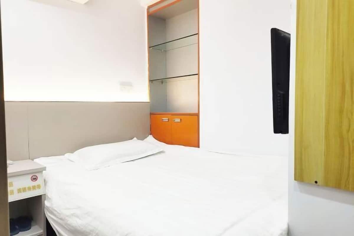 Habitación de hotel con cama, armario con varios estantes, mesita de noche y televisor montado en la pared.