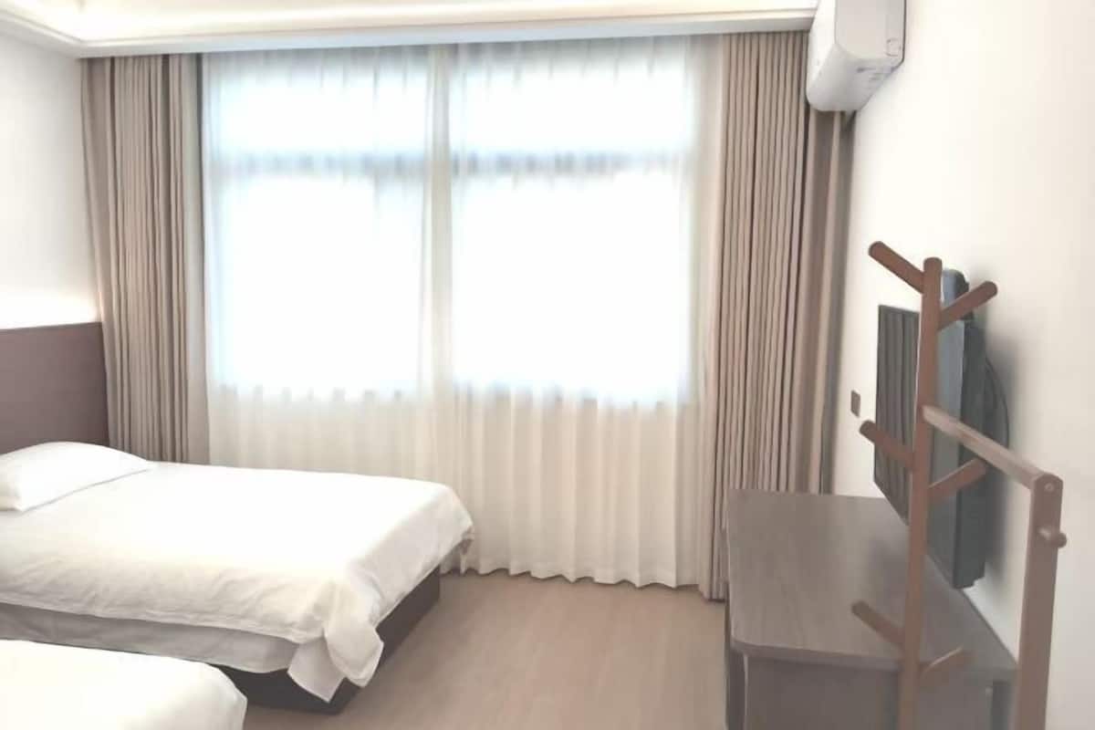Una habitación de hotel con dos camas, una silla de madera, un perchero y un ventanal con cortinas.