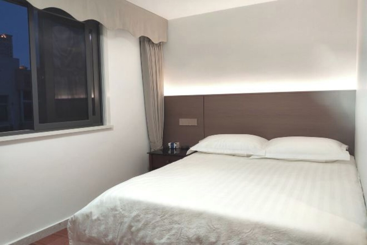 A hotel room with a bed, a nightstand, a window with curtains, and a wall-mounted light.