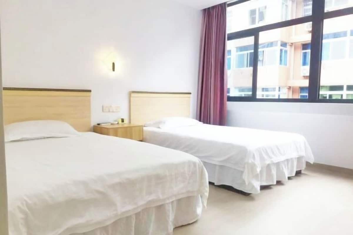 Habitación de hotel con dos camas, una mesita de noche, una lámpara de pared y una ventana con vistas a edificios.