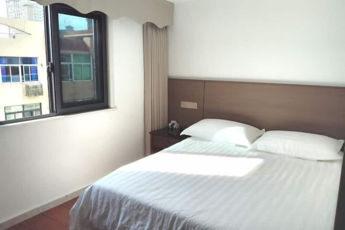 Una habitación de hotel con cama, mesita de noche, ventana con cortinas y vistas a edificios.