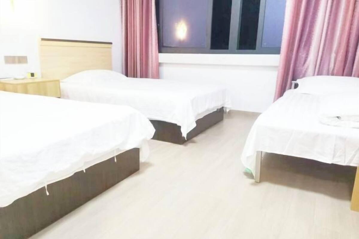Una habitación de hotel con tres camas individuales, una mesita de noche con lámpara y una ventana con cortinas.