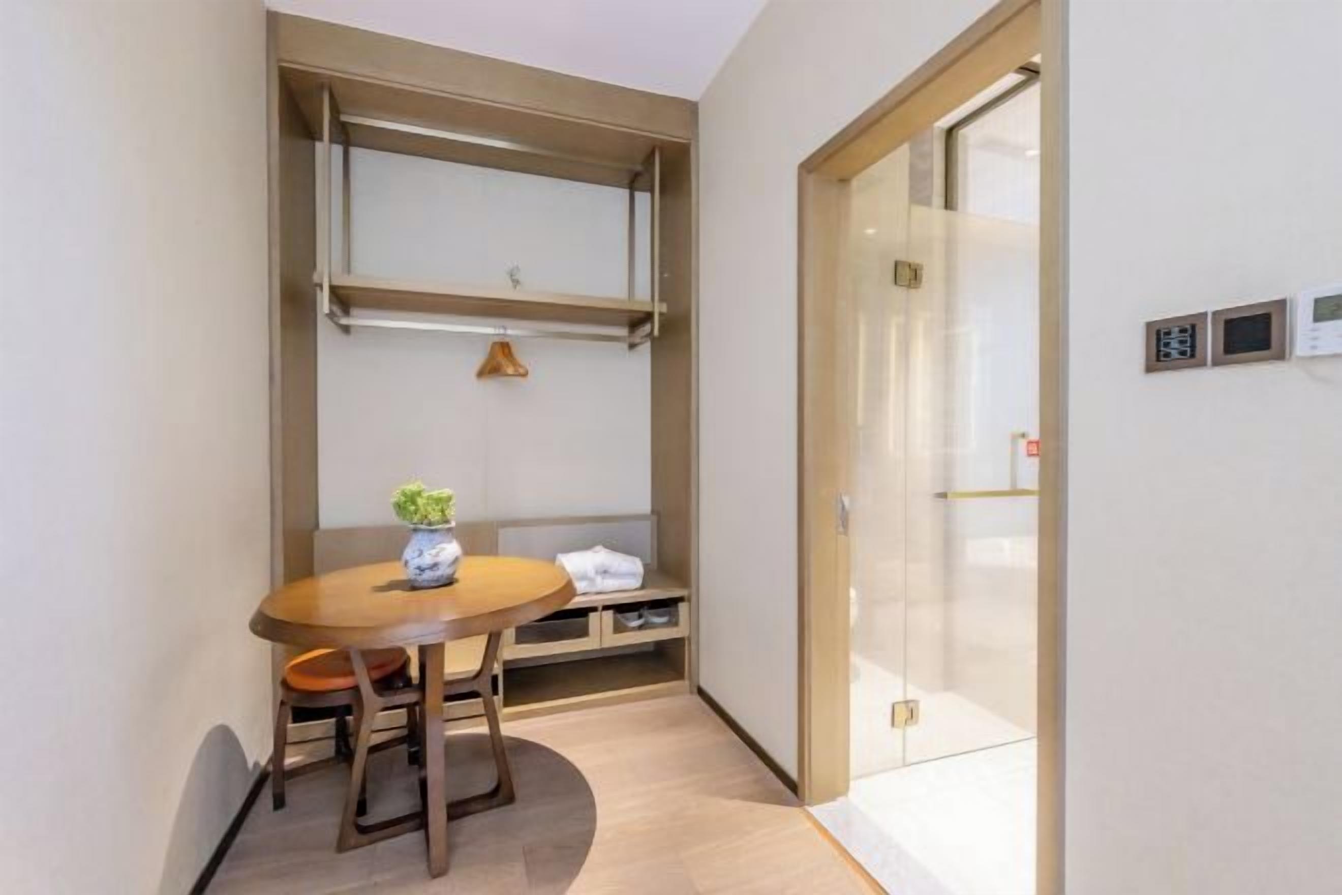 A compact room with a wooden table, a bench, and a small potted plant. There is a sliding door with a glass panel and a wall-mounted control panel.