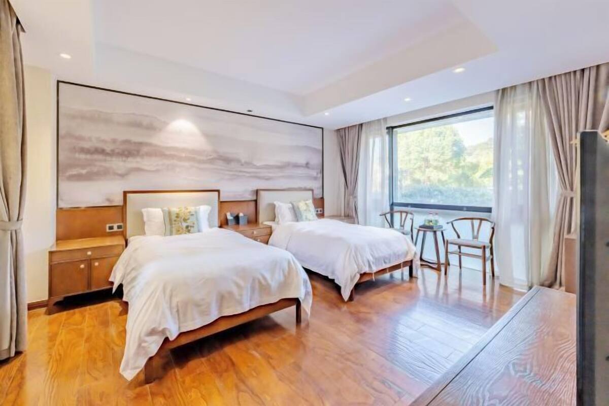 A modern hotel room with two beds, a large window with curtains, a wooden floor, and a wall-mounted artwork.