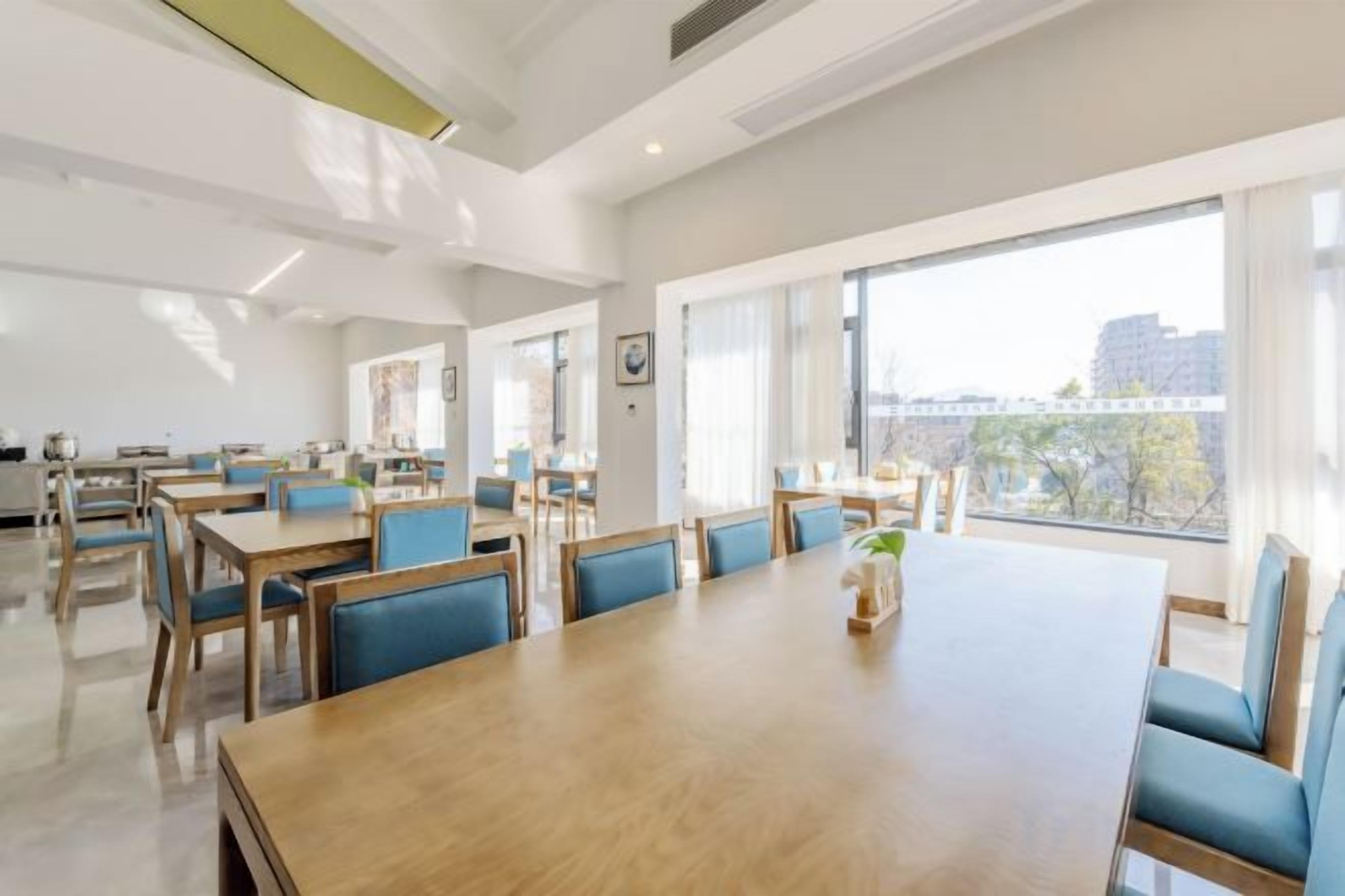 A spacious dining area with a long wooden table and blue upholstered chairs, large windows with a view of buildings, and a modern interior design.