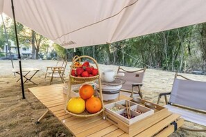 Un montaje para picnic en una playa arenosa con una mesa de madera, una cesta de mimbre con frutas y un tetera blanca bajo una toldo.