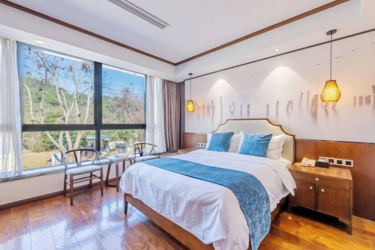 A bedroom with a large window, a bed, a bedside table, and a chair.