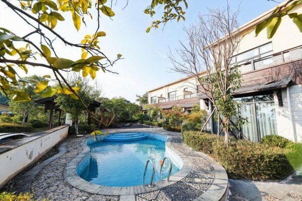A swimming pool with a tiled deck, surrounded by greenery and a building in the background.