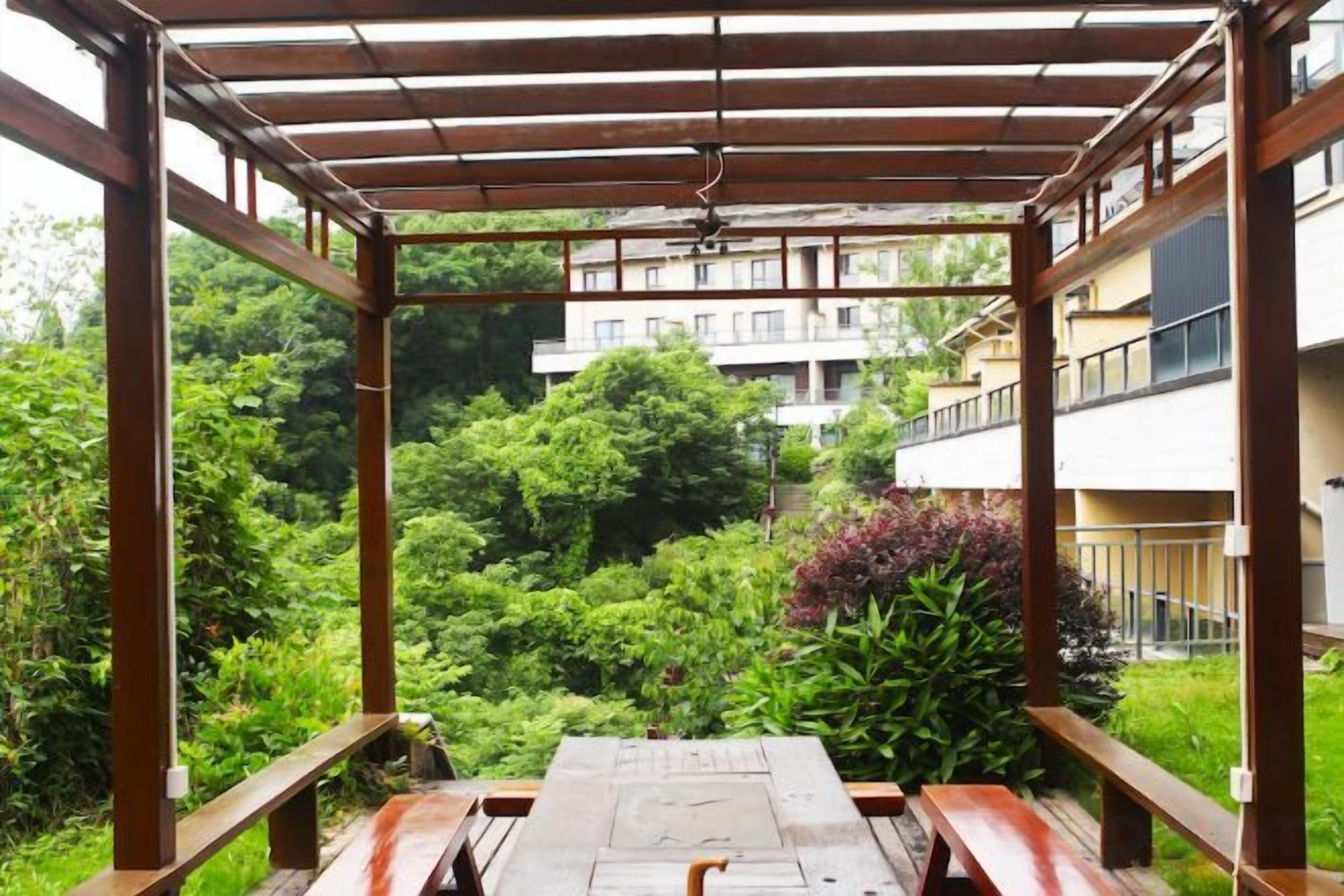 Un espacio al aire libre cubierto con un banco de madera, rodeado de abundante vegetación y vistas a un edificio residencial.