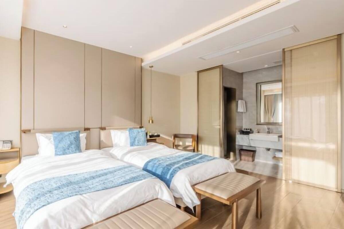A modern hotel room with two beds, a small desk, a bench, and a bathroom visible through an open door.